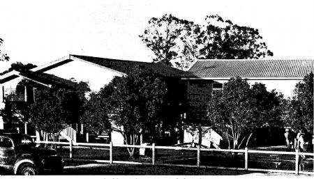 Black and white historic photo of a school building with trees, fencing, and early vehicles in front.